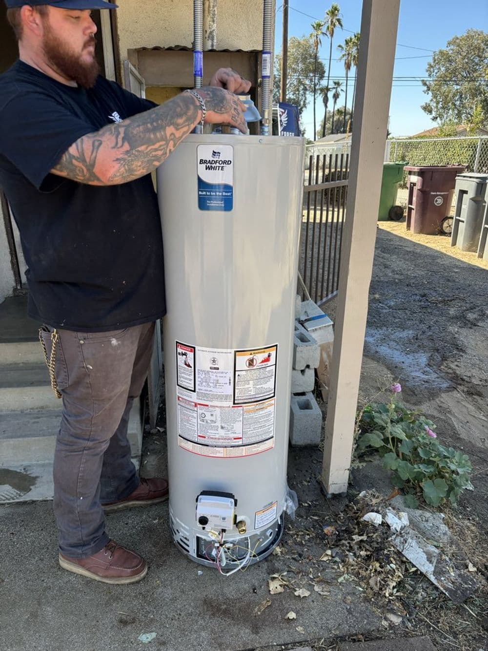 Technician installing a water heater outdoors beside trash bins and plants.