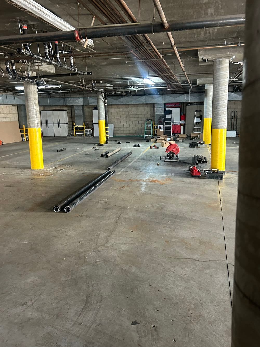 Construction site interior with exposed pipes, yellow safety columns, and tools scattered on the floor.