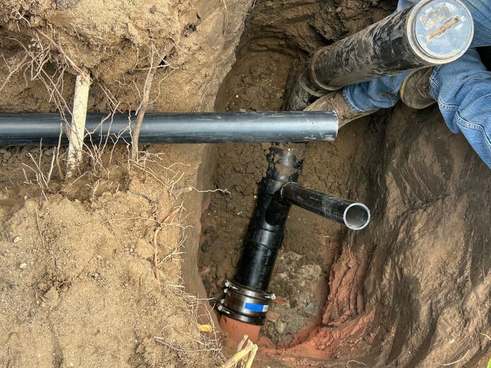 Worker repairing underground plumbing with black pipes in a dirt trench.