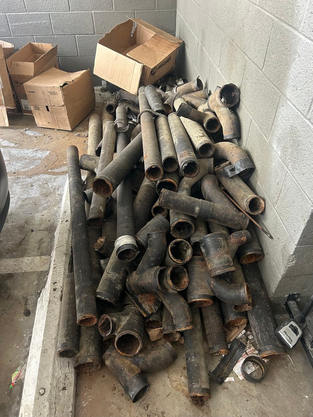 Pile of old, dirty pipes and fittings near a concrete wall in a storage area.