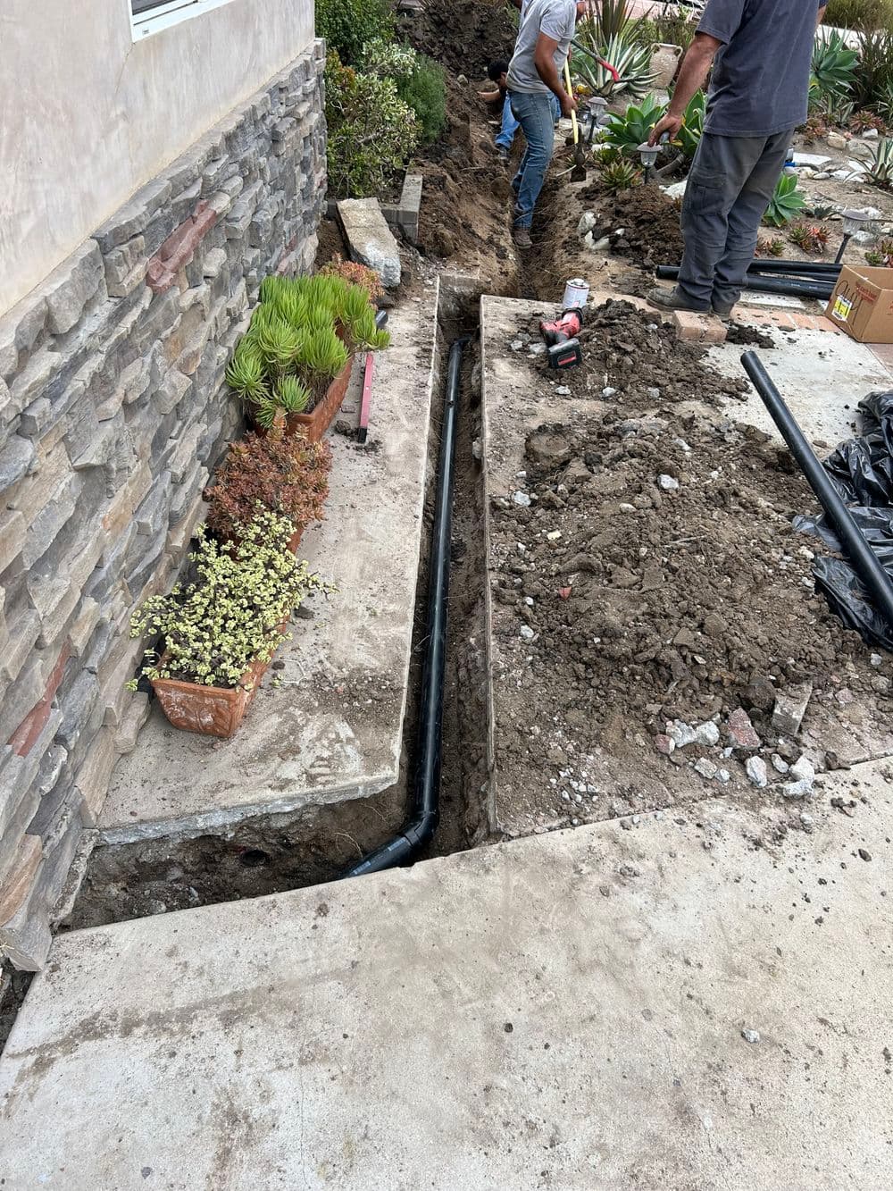 Workers digging a trench for new drainage system beside a house with landscaping.