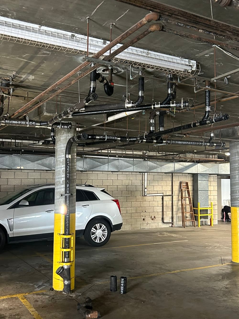 Pipes and supports in a parking garage with a parked SUV and ladder in the background.
